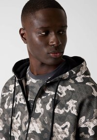 Felpa con cappuccio grigia mimetica con zip e design a motivo bianco, caratterizzata da coulisse nere e finitura testurizzata. Presenta un logo sul petto.