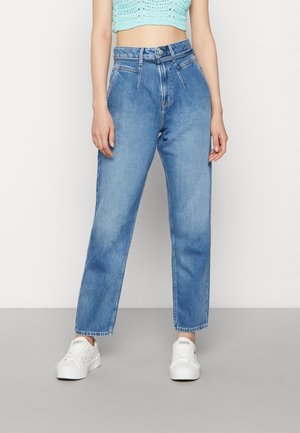 Femme portant un jean taille haute bleu, des baskets blanches à plateforme et un crop top en crochet bleu clair, debout devant un fond uni.