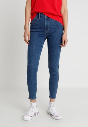 Persoon draagt hoog getailleerde blauwe skinny jeans met ritsen bij de enkels, witte hoge sneakers en een rood mouwloos shirt.
