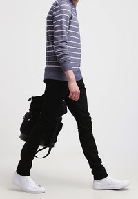 Persoon die loopt met zwarte broek, witte sneakers en een blauw gestreept trui, terwijl hij een zwarte rugzak in zijn linkerover hand draagt.