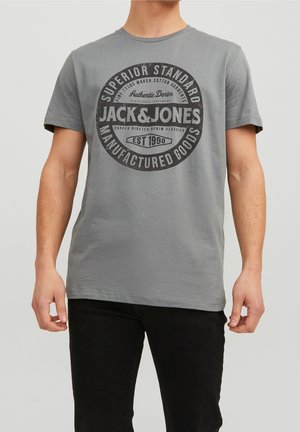 Mężczyzna noszący szarą koszulkę Jack & Jones z czarnym napisem i okrągłym logo, w połączeniu z czarnymi spodniami, stojący na jednolitym tle.