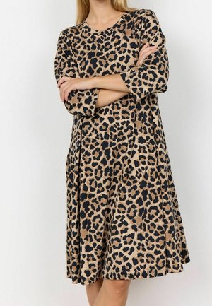Kvinde iført knælængde, langærmet leopardprint kjole med armene krydset mod en ensfarvet lys baggrund.