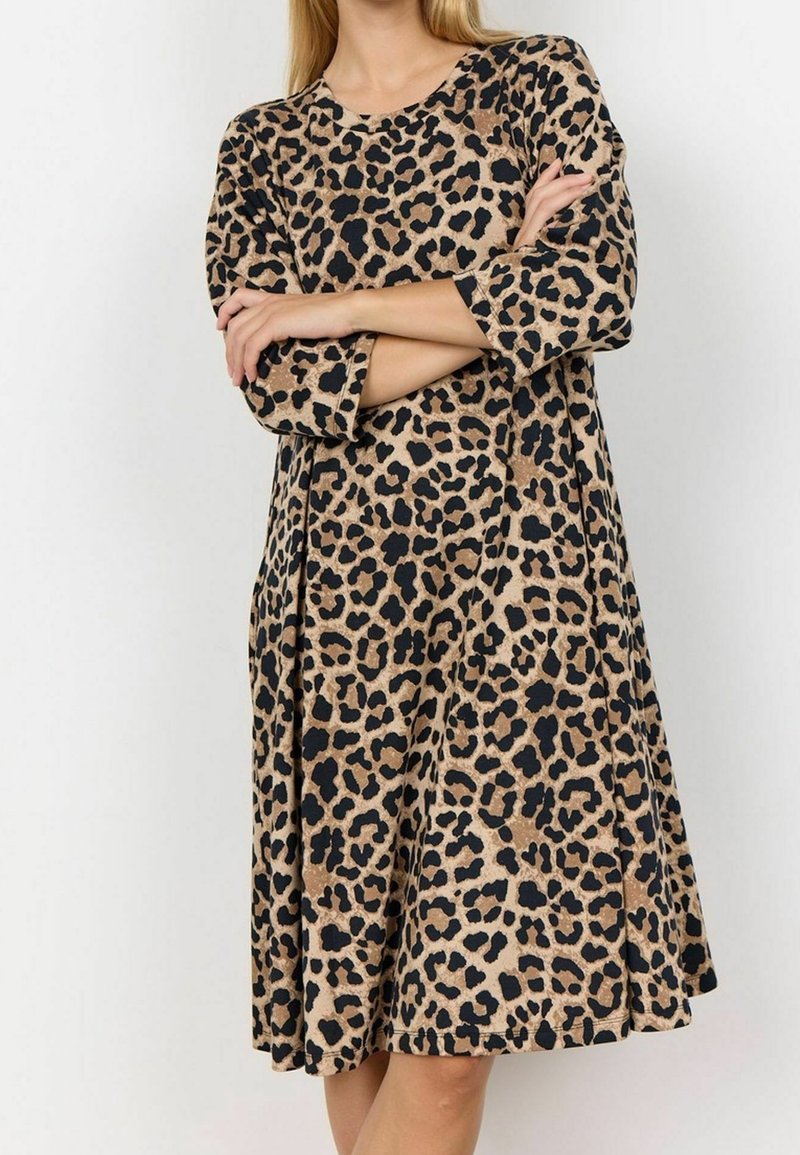 Femme portant une robe imprimée léopard à manches longues et longueur genou, bras croisés devant un fond clair uni.