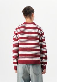 Strikkegenser med maroon og hvite striper, kragehals og ribbestrikket kant. Båret med lyseblå denim, viser en avslappet passform.