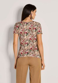 Frau in einem kurzärmligen Top mit floralem und Leopardenmuster und beigen Hosen, die von hinten zeigt. Die Rückendetails sind sichtbar, ebenso wie lila statement Ohrringe.
