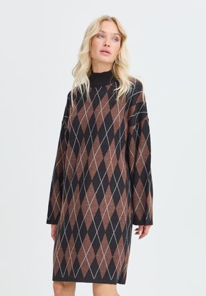 Femme blonde portant une robe-pull à manches longues noire et marron à motif losanges argyle avec un col montant, debout devant un fond uni.