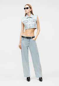 Kobieta w czarnych okularach przeciwsłonecznych, skróconej jeansowej kamizelce bez rękawów, szerokich jasnoniebieskich dżinsach, czarnym pasku i czarnych szpiczastych botkach, stojąca.