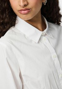 Camicia bianca con colletto classico, caratterizzata da una sottile trama e bottoni bianchi lucidi, mostrata parzialmente su un modello.