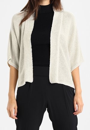 Woman wearing a loose, light beige knit cardigan over a fitted black ribbed turtleneck and black high-waisted pants.