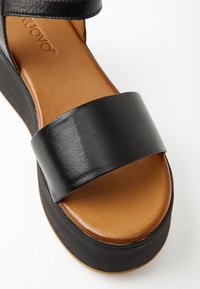 Černé kožené platformové sandály s širokým předním páskem, hnědým polstrovaným stélkou a černou podrážkou na bílém pozadí.