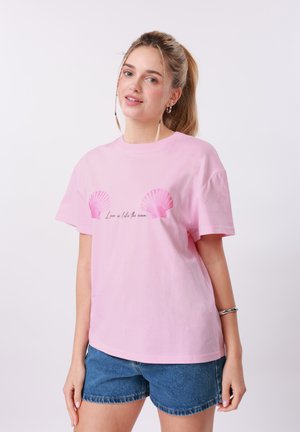 Noor naine blondi hobusesabaga, kes kannab roosat T-särki roosade karpide ja kirjaga ning siniseid teksapükse, seistes valge tausta ees.