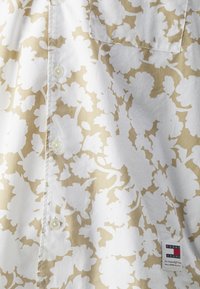 Camicia con motivo floreale in bianco e beige, realizzata in tessuto leggero. Presenta bottoni e una tasca prominente, con un'etichetta di marca in basso.