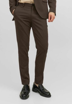 Uomo che indossa pantaloni marroni su misura con mocassini neri in pelle, camicia beige e blazer marrone abbinato, in piedi su uno sfondo semplice.