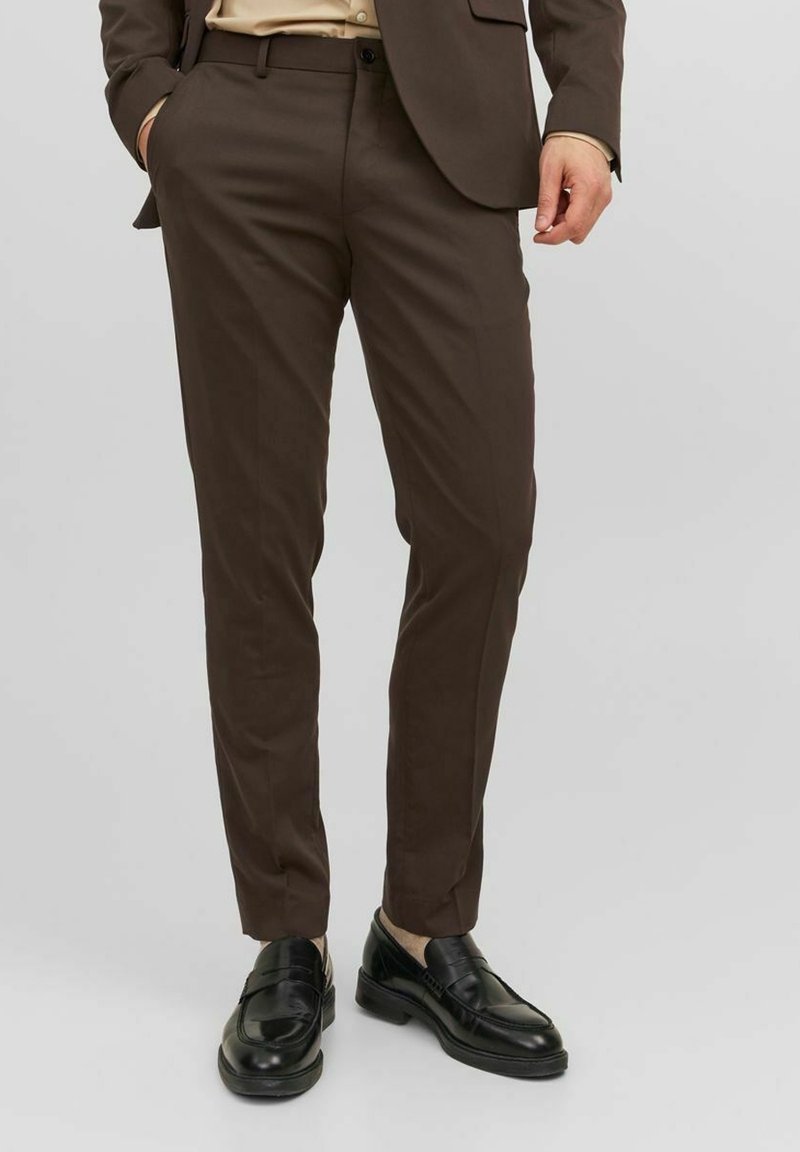 Homme portant un pantalon marron sur mesure avec des mocassins en cuir noir, une chemise beige et une veste marron assortie, debout devant un fond uni.