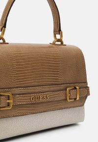 Brązowa torebka skórzana o teksturowanej powierzchni z akcentami w postaci złotych klamerek oraz wytłoczonym logo "GUESS" na przednim pasku, umieszczona na jasnobeżowej tkaninie.