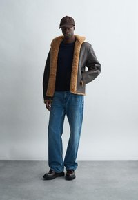 Ruskea nahkatakki, jossa on pörröinen vaaleanruskea kaulus, päällä tummansinisellä paidalla; yhdistetty vaaleansiinisiin farkkuihin ja tummiin kenkiin.