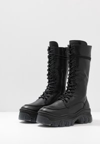 Bottes en cuir noir montant jusqu'aux genoux avec une semelle épaisse, un laçage à l'avant et une fermeture éclair latérale pour un enfilage facile. Détails texturés sur la semelle.