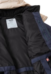 Izolowana kurtka z beżową zewnętrzną warstwą, czarną podszewką, granatowymi akcentami i kieszenią na zamek. Metka wewnętrzna pokazuje markę i rozmiar.