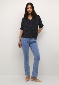 Blusa nera a maniche corte con pois bianchi, con bottoni sul davanti e vestibilità rilassata. Abbinata a jeans a zampa d'elefante di colore blu chiaro e décolleté beige.