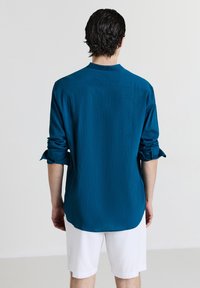 Türkises Langarm-Shirt aus strukturiertem Stoff, mit lockerer Passform und hochgekrempelten Ärmeln, kombiniert mit weißen Shorts, von hinten betrachtet.