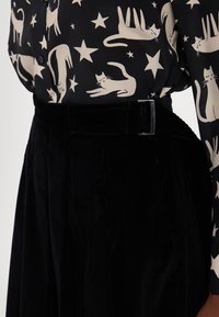 Czarne spodnie z sztruksu z wysokim stanem i szerokimi nogawkami, zestawione z wzorzystą czarną bluzką ozdobioną białymi wzorami kotów i gwiazd.