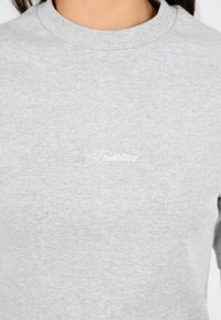 Primo piano di una persona che indossa un maglione a costine grigio chiaro con del piccolo testo ricamato bianco centrato sul petto.