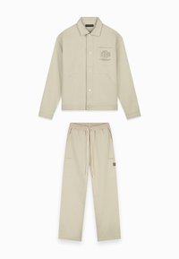Beige jas met een knoopsluiting, voorkruizen en een logo op de zak; gecombineerd met beige sweatpants met een elastische tailleband en zijzakken.