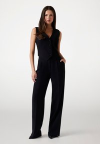 Black sleeveless vest with a fitted design and five buttons, paired with high-waisted, wide-leg black trousers featuring side pockets.