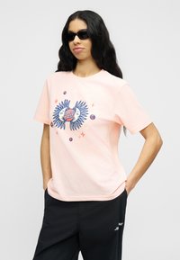 Νεαρός με μακριά σκούρα μαλλιά που φοράει μαύρα γυαλιά ηλίου, ροζ ανοιχτό t-shirt με σχέδιο ήλιου και φεγγαριού, και μαύρο παντελόνι με τα χέρια στις τσέπες.