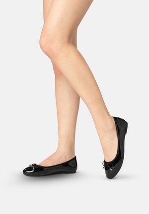 Blote benen met glanzende zwarte ballerina’s met kleine strikaccenten, één voet plat en de andere iets verhoogd op een witte achtergrond.