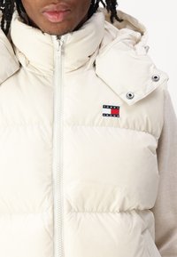 Bež prsluk s pufanjem i visokim ovratnikom, prednjim patent zatvaračem, kopčama na dugmad i malim "Tommy Jeans" logotipom na lijevoj strani grudi. Glatka, sjajna tekstura.
