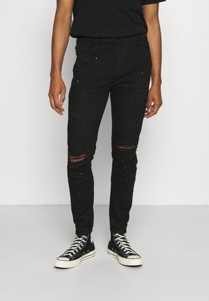 Jeans de mezclilla negra con áreas desgastadas y detalles salpicados. Corte ajustado con una cintura de altura media. Combinados con zapatillas altas negras.