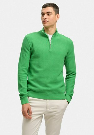 Grøn kvart-zip trøje, struktureret strikstof, tætsiddende design, ribbede manchetter og kant, parret med lyse bukser.