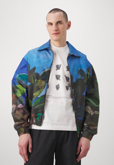 Barevná bomber bunda s modrým a zeleným květinovým potiskem, zapínání na zip v přední části a elastickými manžetami. Nositá přes bílý tričko s grafickými detaily.