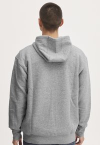 Man with short hair wearing a plain gray hoodie, facing away, against a neutral light background.