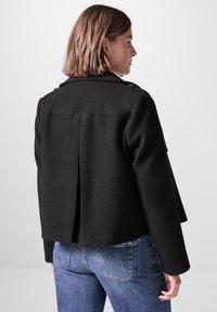 Svart jacka med skräddarsydd passform, strukturerade axlar och subtilt veckdetalj på baksidan. Tyget verkar slätt och texturerat.