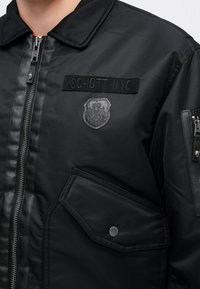 Veste noire avec fermeture éclair garnie de cuir, écusson indiquant "SCHOTT NYC,", un emblème en forme de bouclier, et une poche à pression à l'avant.