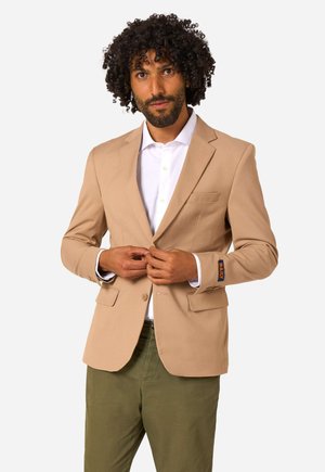 Mann mit lockigem Haar, der einen beigefarbenen Blazer über einem weißen Hemd zuknöpft, trägt olivgrüne Hosen, vor einfachem hellem Hintergrund.