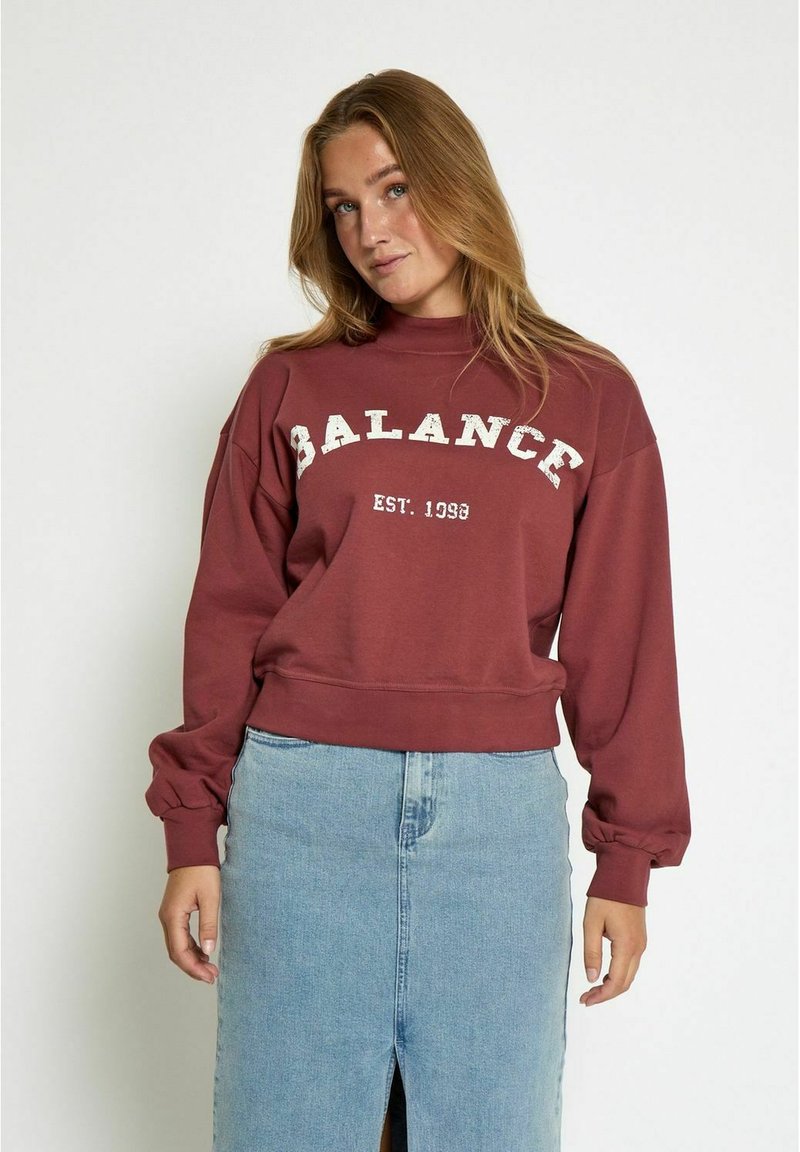 Sudadera color burdeos con "BALANCE EST. 1998" en blanco, con cuello alto, hombros caídos y puños acanalados. Combinada con una falda de mezclilla.