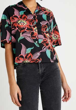 Osoba nosząca czarną koszulę z krótkim rękawem zapinaną na guziki, ozdobioną dużymi czerwono-różowymi kwiatowymi wzorami, oraz ciemnoszare jeansy z wysokim stanem.