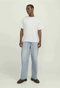 Man staat gekleed in een wit oversized t-shirt, lichtblauwe wijde jeans en donkerbruine schoenen tegen een effen lichte achtergrond.