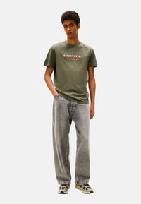 T-shirt in cotone verde oliva con grafica "tommy jeans", abbinato a jeans in denim grigio larghi e sneakers con dettagli verdi.