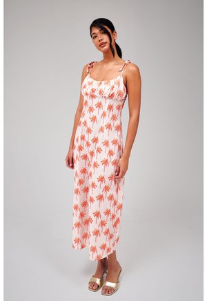 Kobieta stoi, ubrana w białą midi sukienkę bez rękawów z pomarańczowym nadrukiem palm i złote sandały na otwartym palcu na jednolitym szarym tle.