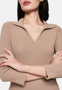 Beiges, geripptes Strickoberteil mit V-Ausschnitt und Kragen. Mit langen Ärmeln und Rüschenabschlüssen. Glatte Textur, figurbetonte Silhouette.