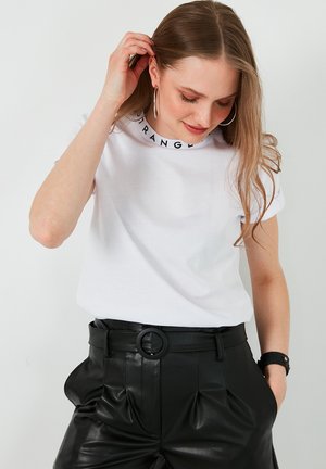 Camiseta blanca de algodón con un cuello impreso en negro que dice "STRANGE". Combinada con pantalones de cuero negros de tiro alto que tienen un cinturón.