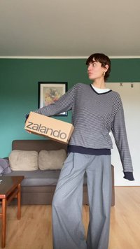 Gestreepte gebreide trui in marineblauw en wit, gecombineerd met wijde grijze broek. Houdt een Zalando-karton vast, staande in een woonkamer.
