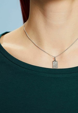 Zilveren ketting met een rechthoekige hanger met een reliëfpatroon, gedragen over een groene blouse, die de halslijn accentueert.