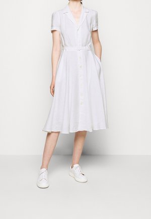 Vrouw draagt een witte midi-jurk met korte mouwen en knopen met een riem, gecombineerd met witte vetersneakers, staand tegen een witte achtergrond.