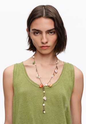 Kobieta o krótkich brązowych włosach ubrana jest w zielony top bez rękawów oraz naszyjnik z pereł i małych zawieszek w kształcie muszelek na białym tle.