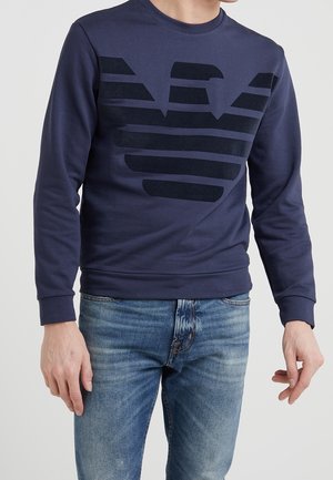 Mann trägt marineblauen Pullover mit großem schwarzem Grafik-Logo auf der Brust, kombiniert mit blauen Jeans, vor neutralem Hintergrund.